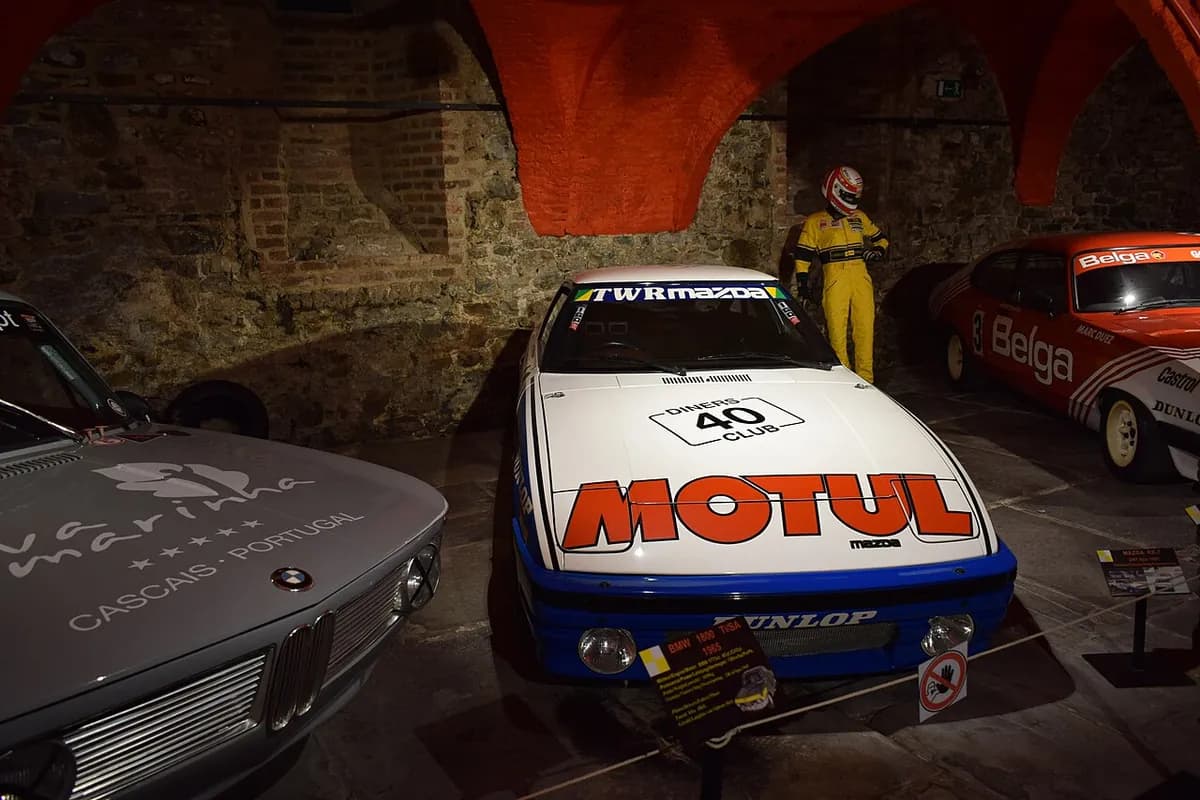 Racing cars displayed under the brick vaults of Stavelot Abbey, including a BMW 1800 TI/SA, a TWR Mazda RX-7 and a Belga Ford Capri