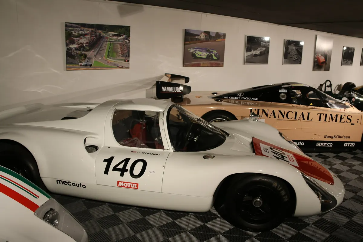 The white Porsche 910, number 140, on display in the Spa-Francorchamps Circuit Museum, seen in profile, with a Porsche 962 in Financial Times livery in the background